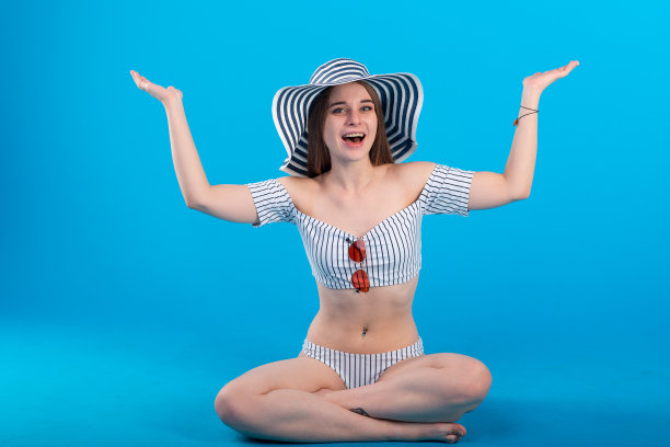 年轻女子举起双手在白色条纹泳装内衣和条纹帽子孤立在蓝色背景图片下载