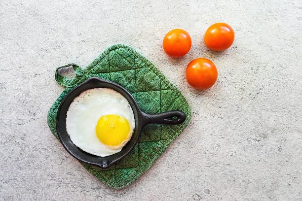 在黑色的铸铁平底锅上煎鸡蛋。在一个黑色的小铁锅里煎鸡蛋。灰色大理石桌上放着三个西红柿。图片下载
