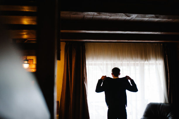 一个英俊自信的年轻人站在白色衬衫和西装外套。英俊的男人站在窗边看向别处。图片下载