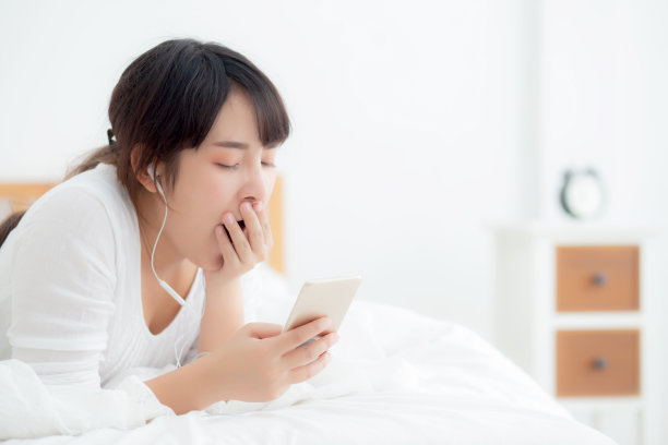 美丽的年轻亚洲女人躺在床上，打着哈欠看智能手机上的社交媒体，亚洲女孩看手机或听音乐昏昏欲睡，女性懒惰和无聊，生活理念。图片下载