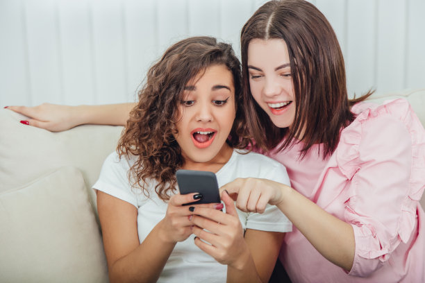 漂亮的年轻女孩坐在沙发上，在家里，看着手机里的照片，微笑着，互动。一个女孩拿着手机，另一个用手指着屏幕。图片下载