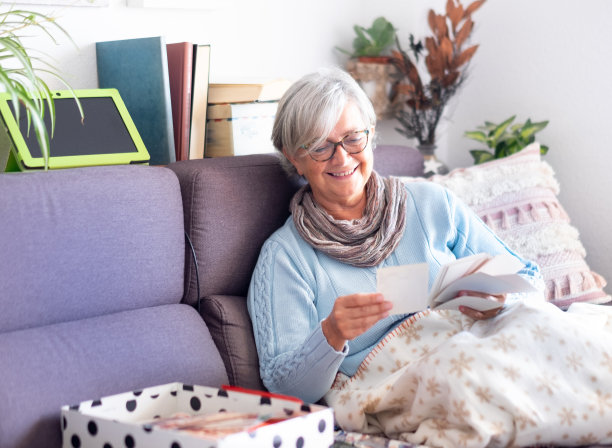 正面的观点，一个有吸引力的资深女士看老照片的一生微笑。坐在沙发上，用手机和平板电脑。白色的墙壁和书的背景图片下载