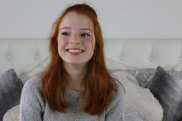 照片中，一个14 / 15岁的红头发、皮肤苍白、长着雀斑的少女坐在卧室里，穿着灰色羊毛套头衫，涂着裸色口红，靠在灰白的靠垫和枕头上，微笑着看着镜头，神情轻松图片下载