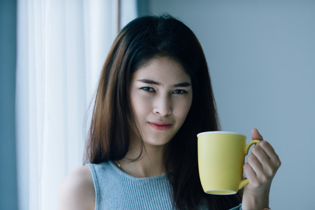 亚洲妇女拿着黄色马克杯靠近窗户与自然光线。图片下载
