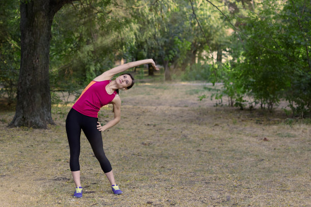 美丽的年轻苗条的女人使健身房在早晨公园。倾斜下来，明亮的运动服，白色耳机，冷静和放松气氛，伸展训练。图片下载