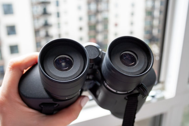 一个女人的手拿着黑色的双筒望远镜在一个模糊的背景窗户与一个邻近的房子观察邻居，环境或自然的特写视图图片下载