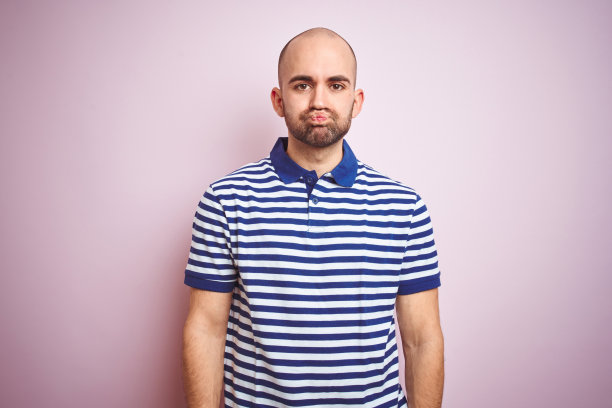 年轻的秃顶男子与胡子穿着休闲条纹蓝色t恤在粉红色孤立的背景蓬松面颊与滑稽的脸。嘴巴里充满了气，疯狂的表情。图片下载