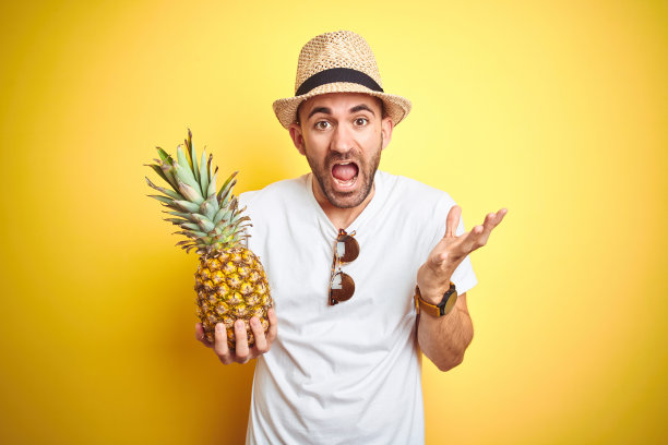 年轻人戴着夏季帽子，拿着菠萝在黄色的背景非常高兴和兴奋，胜利者的表情庆祝胜利尖叫着大大的微笑和举起手图片下载