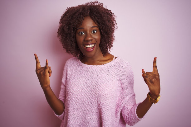 年轻的非洲黑人妇女穿着毛衣站在孤立的粉红色背景大喊与疯狂的表情做摇滚符号与手举起。音乐明星。沉重的概念。图片下载