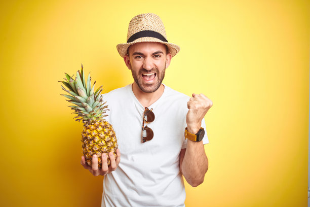 年轻人戴着夏帽，拿着菠萝在黄色的背景下尖叫着自豪，庆祝胜利和成功，非常激动，欢呼的情绪图片下载