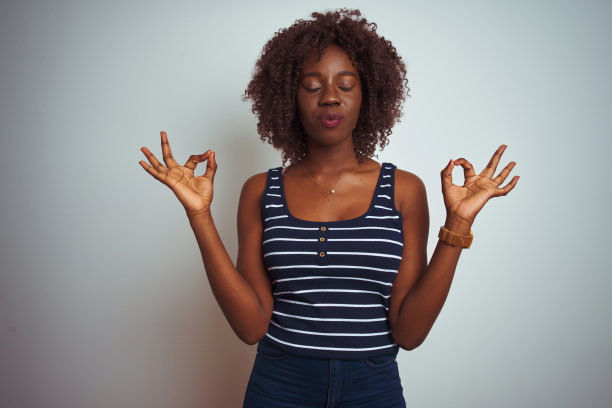 年轻的非洲黑人妇女穿着条纹t恤站在孤立的白色背景放松和微笑，闭上眼睛做冥想手势与手指。瑜伽的概念。图片下载