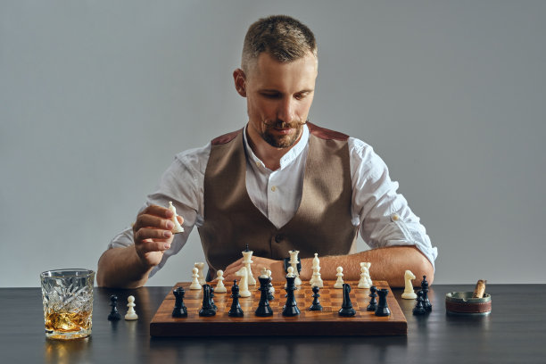 男人留着时髦的胡子，穿着棕色的背心，白色的衬衫正在下棋。旁边放着一杯威士忌，烟灰缸，雪茄。灰色背景,特写图片下载
