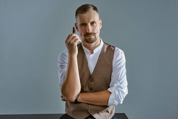 一个留着时髦胡子的男人，穿着棕色背心和白色衬衫，正坐在一张桌子上，享受着雪茄。灰色背景,特写图片下载