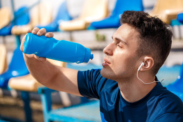 年轻男子戴着耳机在体育场外面坐着听音乐，喝着水图片下载