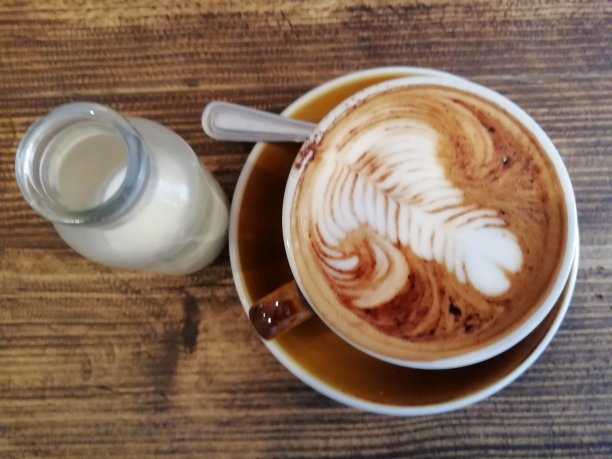 卡布奇诺在一个棕色的杯子和茶托，勺子和一小瓶牛奶放在一张木桌上图片下载