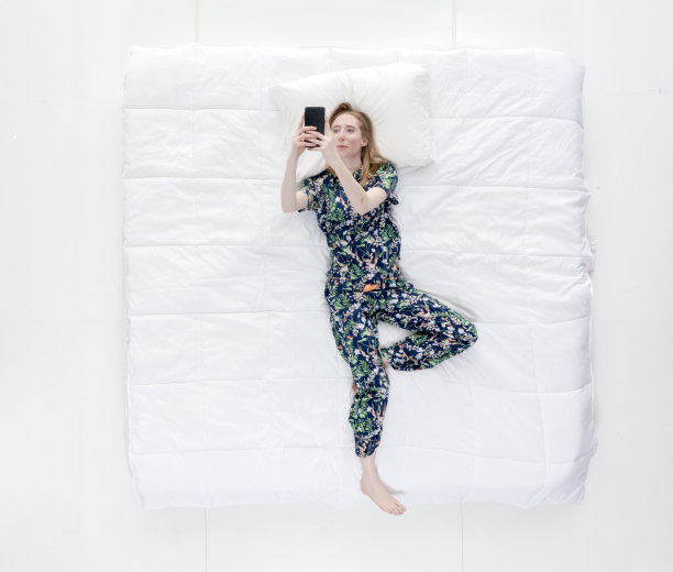 上图/全身/一个20-29岁的成年美丽白人女性/年轻女性穿着睡衣躺在卧室的白色背景前，拿着手机/使用智能手机图片下载