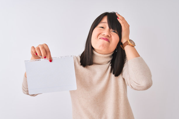 年轻美丽的中国女人拿着笔记本在孤立的白色背景强调与手在头上，震惊与羞愧和惊讶的脸，愤怒和沮丧。为错误而恐惧和不安。图片下载