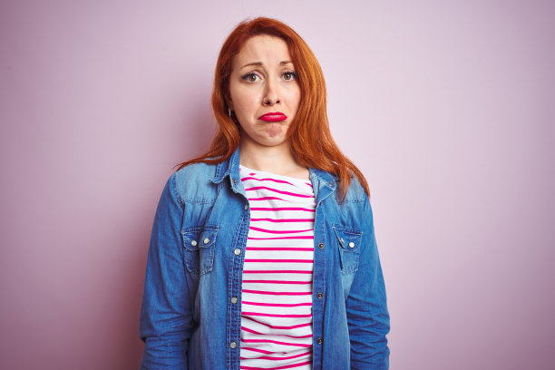 漂亮的红发女人穿着牛仔衬衫和条纹t恤在孤立的粉红色背景沮丧和担心的痛苦，哭泣愤怒和害怕。悲伤的表情。图片下载