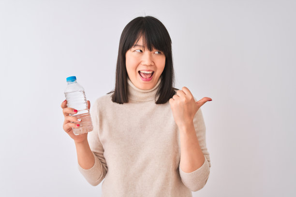 年轻美丽的中国女人拿着水瓶在孤立的白色背景指向和显示拇指向上到一边与幸福的脸微笑图片下载