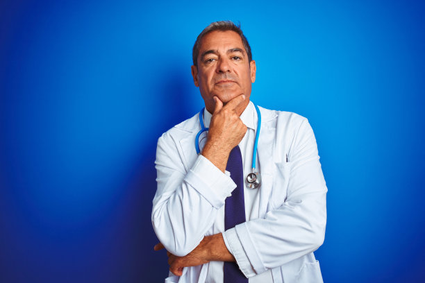 英俊的中年医生男子戴着听诊器在孤立的蓝色背景看起来自信在镜头微笑交叉的胳膊和手高举在下巴。积极的思考。图片下载
