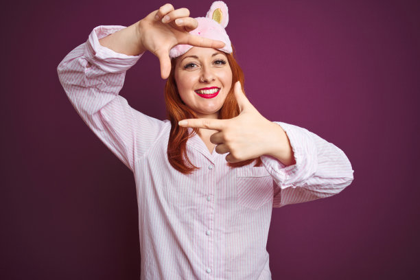 年轻美丽的红头发女人穿着睡衣和面具在紫色孤立的背景微笑与手和手指框架与快乐的脸。创意和摄影概念。图片下载