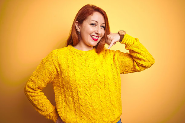 美丽的红头发女人穿着冬季毛衣站在孤立的黄色背景微笑做电话手势的手和手指，就像在讲电话。沟通的概念。图片下载