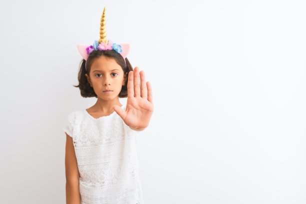 美丽的小女孩戴着独角兽冠冕站在孤立的白色背景上用手掌做着停止歌唱。警告表情与否定和严肃的手势在脸上。图片下载