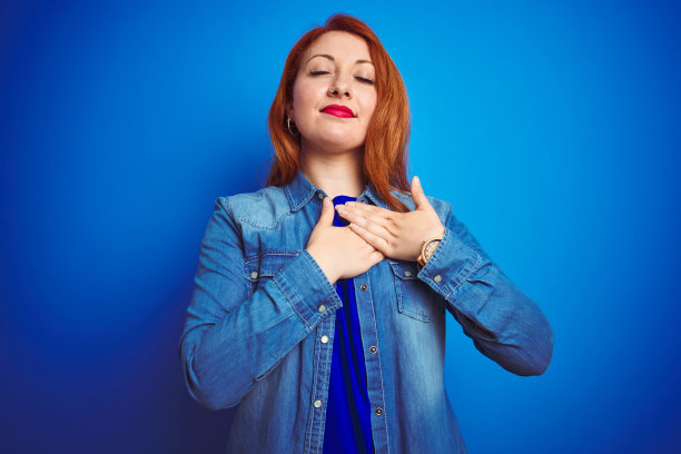 年轻美丽的红头发女人穿着牛仔衬衫站在蓝色孤立的背景微笑与双手在胸前闭上眼睛和感激的手势在脸上。健康的概念。图片下载