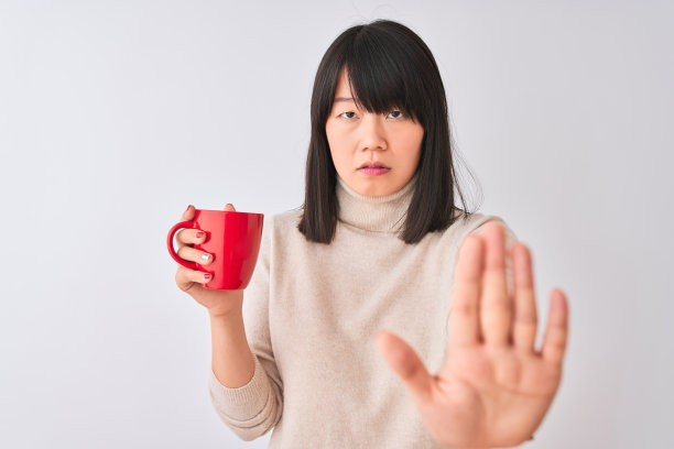 年轻美丽的中国女人喝红杯咖啡在孤立的白色背景与开放的手做停止标志与严肃和自信的表情，防御手势图片下载