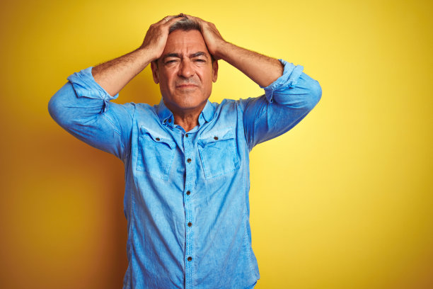 英俊的中年男子穿着牛仔衬衫站在孤立的黄色背景遭受头痛绝望和压力，因为疼痛和偏头痛。手放在头上。图片下载
