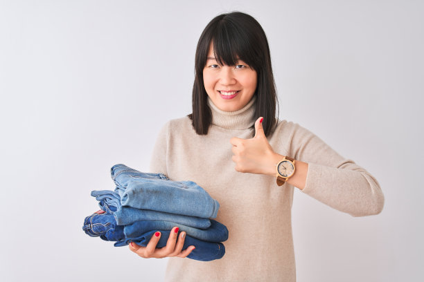 美丽的中国女店主拿着折叠的牛仔裤在孤立的白色背景愉快地与大微笑做ok的手势，拇指向上的手指，优秀的手势图片下载