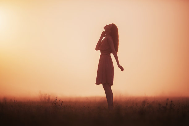 在黎明的背景下，美丽的红发女孩穿着粉红色的裙子。女孩奔向日出。雾蒙蒙的晨曦笼罩着田野。黄色的夏季日出。明亮的朝阳。女性的身影图片下载