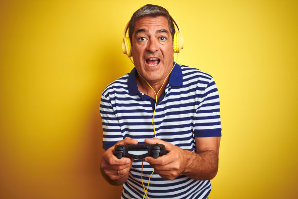 中年男子玩电子游戏，使用耳机在孤立的黄色背景吓了一跳，吃惊的脸，害怕和兴奋的恐惧表情图片下载