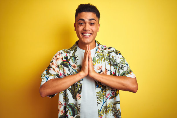 年轻的巴西男子度假穿着夏天的花衬衫在孤立的黄色背景祈祷与手一起请求原谅微笑自信。图片下载
