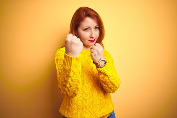 漂亮的红发女人穿着冬季毛衣站在孤立的黄色背景准备打架用拳头防御手势，愤怒和不安的脸，害怕问题图片下载