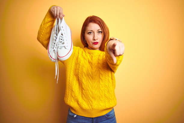 年轻美丽的红发女人拿着白色运动鞋在黄色孤立的背景指向指向相机和你，手势，积极和自信的手势从前面图片下载