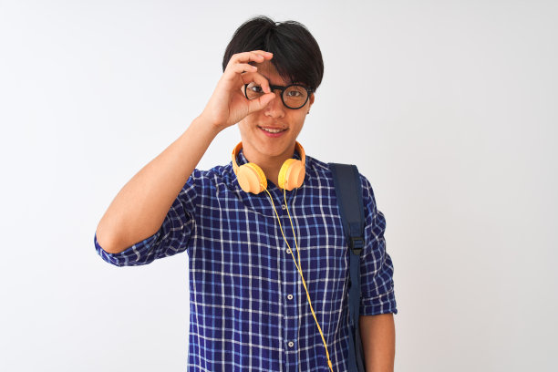 中国学生男子背着背包，戴着耳机，在孤立的白色背景下做着ok的手势，手带微笑，眼睛透过手指看着幸福的脸。图片下载