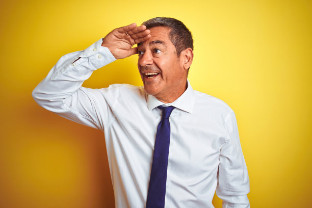 英俊的中年商人站在孤立的黄色背景非常高兴，微笑着看远远的手在头上。搜索概念。图片下载