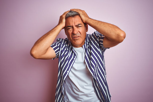 英俊的中年男子穿着条纹衬衫站在孤立的粉红色背景下遭受头痛绝望和压力，因为疼痛和偏头痛。手放在头上。图片下载