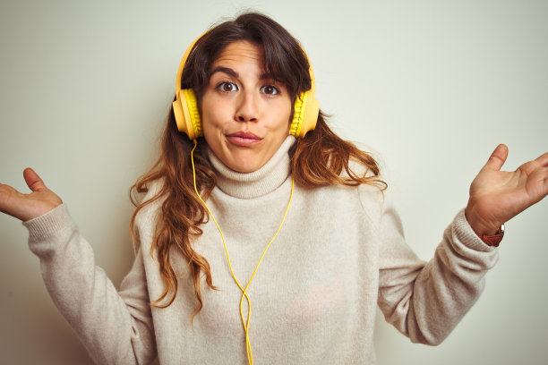 年轻美丽的女人听音乐使用耳机在白色孤立的背景下，愚蠢和困惑的表情与手臂和手举起。怀疑的概念。图片下载