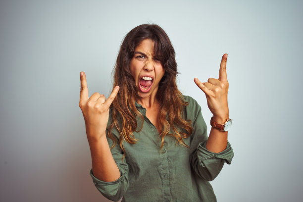 年轻美丽的女人穿着绿色衬衫站在灰色孤立的背景大喊与疯狂的表情做摇滚符号与手举起。音乐明星。沉重的音乐概念。图片下载