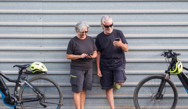 两辆电动自行车和两个白发老人在看手机。黑色的衣服和太阳镜。在灰色的金属面板后面图片下载