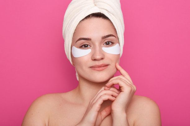 近距离拍摄美丽的年轻女子与胶原蛋白垫下她的眼睛，站在头上戴白色毛巾，迷人温柔的女性保持双手靠近她的脸。Spa，眼部肌肤护理理念。图片下载