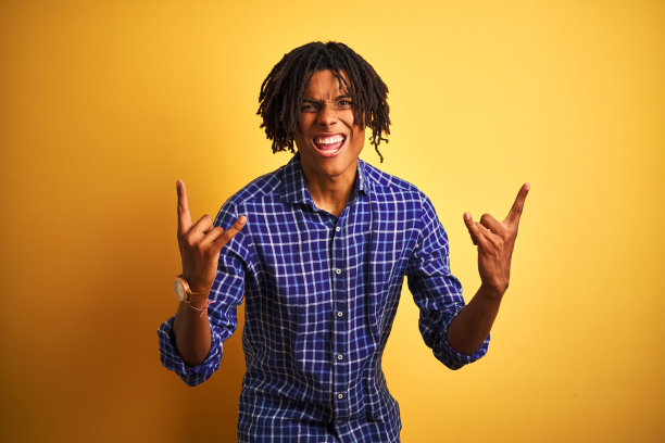 非洲人与雷鬼发穿着休闲衬衫站在孤立的黄色背景大喊与疯狂的表情做摇滚符号与手举起。音乐明星。沉重的概念。图片下载