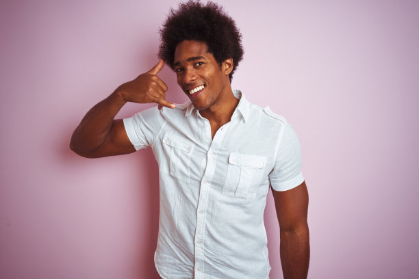 年轻的美国男子与非洲式头发穿白衬衫站在孤立的粉红色背景微笑做电话手势与手和手指，就像在讲电话。沟通的概念。图片下载