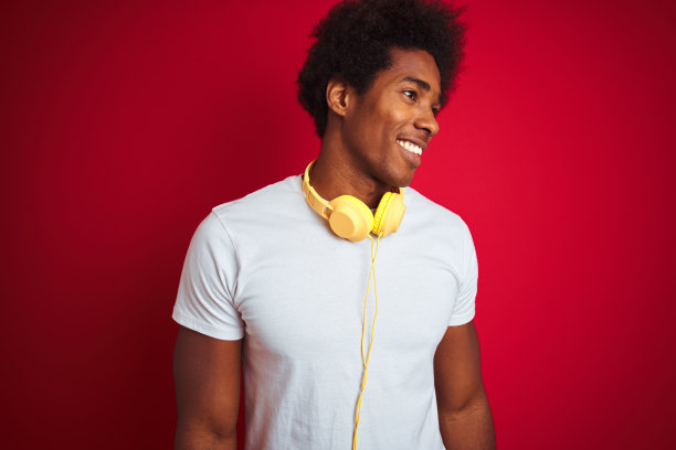 年轻的非洲裔美国人听音乐使用耳机孤立的红色背景看向一边，脸上微笑，自然的表情。自信的笑。图片下载