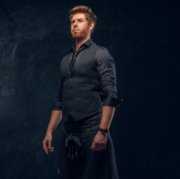 一名红发男子身穿优雅的背心、领带和苏格兰短裙，背靠深色纹理的墙壁图片下载