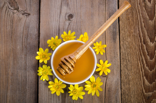 玻璃碗里放着蜂蜜，木制的背景上环绕着黄色的花朵图片下载