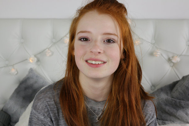 照片中，14 / 15岁的红头发、皮肤苍白、长着雀斑的少女坐在卧室里，穿着斑驳的灰色t恤，靠在灰白相间的靠垫和枕头上，靠在白色的床头板上，轻松地微笑着看着镜头图片下载