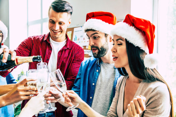 玩得开心!两名男子和一名黑发女子的特写照片，在圣诞老人的帽子拿着长笛和一瓶香槟，与他们的朋友在白色的工作室背景敬酒。图片下载